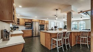 A modern kitchen with wooden cabinets, stainless steel appliances, and a central island with bar stools. - MAINE WATERFRONT + MOUNTAIN COMPOUND • SLEEPS 50! (Bridgton)