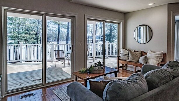 A spacious living room with a large sliding glass door leading to a deck, a comfortable sofa, a coffee table, and a round mirror on the wall. - MAINE WATERFRONT + MOUNTAIN COMPOUND • SLEEPS 50! (Bridgton)
