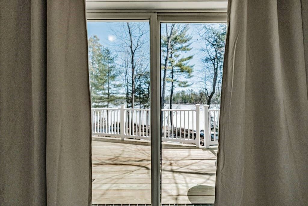 A view through a sliding glass door to an outdoor deck with a white railing and trees in the background.