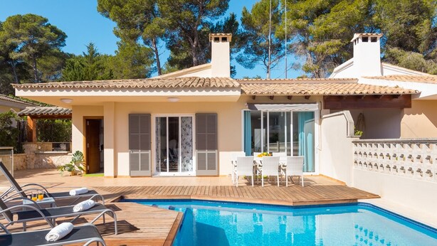 Un espace aménagé au bord de la piscine, avec des chaises longues et une table, une terrasse couverte et une vue sur les arbres environnants.