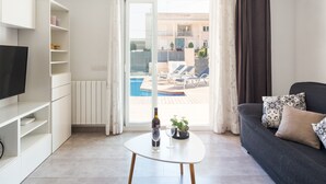 Un salon moderne avec un téléviseur à écran plat, une table basse blanche sur laquelle se trouvent une bouteille et un verre, un canapé foncé agrémenté de coussins à motifs, et une vue sur une piscine et un espace de détente extérieur.