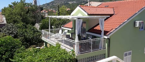 A green house with a red-tiled roof, a balcony with white railings, and a view of a mountain and a church in the background.