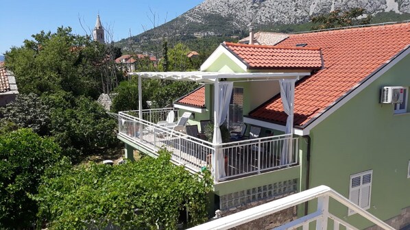 A green house with a red-tiled roof, a balcony with white railings, and a view of a mountain and a church in the background.
