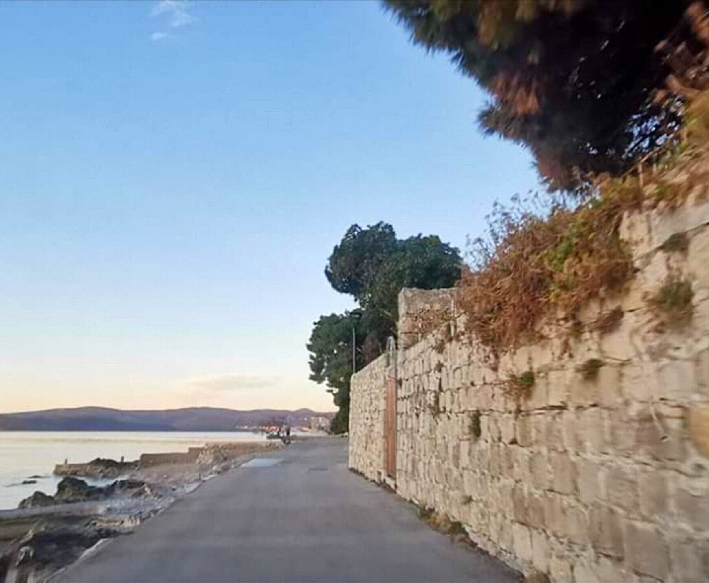 Una carretera pavimentada junto a la costa, con un muro de piedra a un lado y una masa de agua al otro.