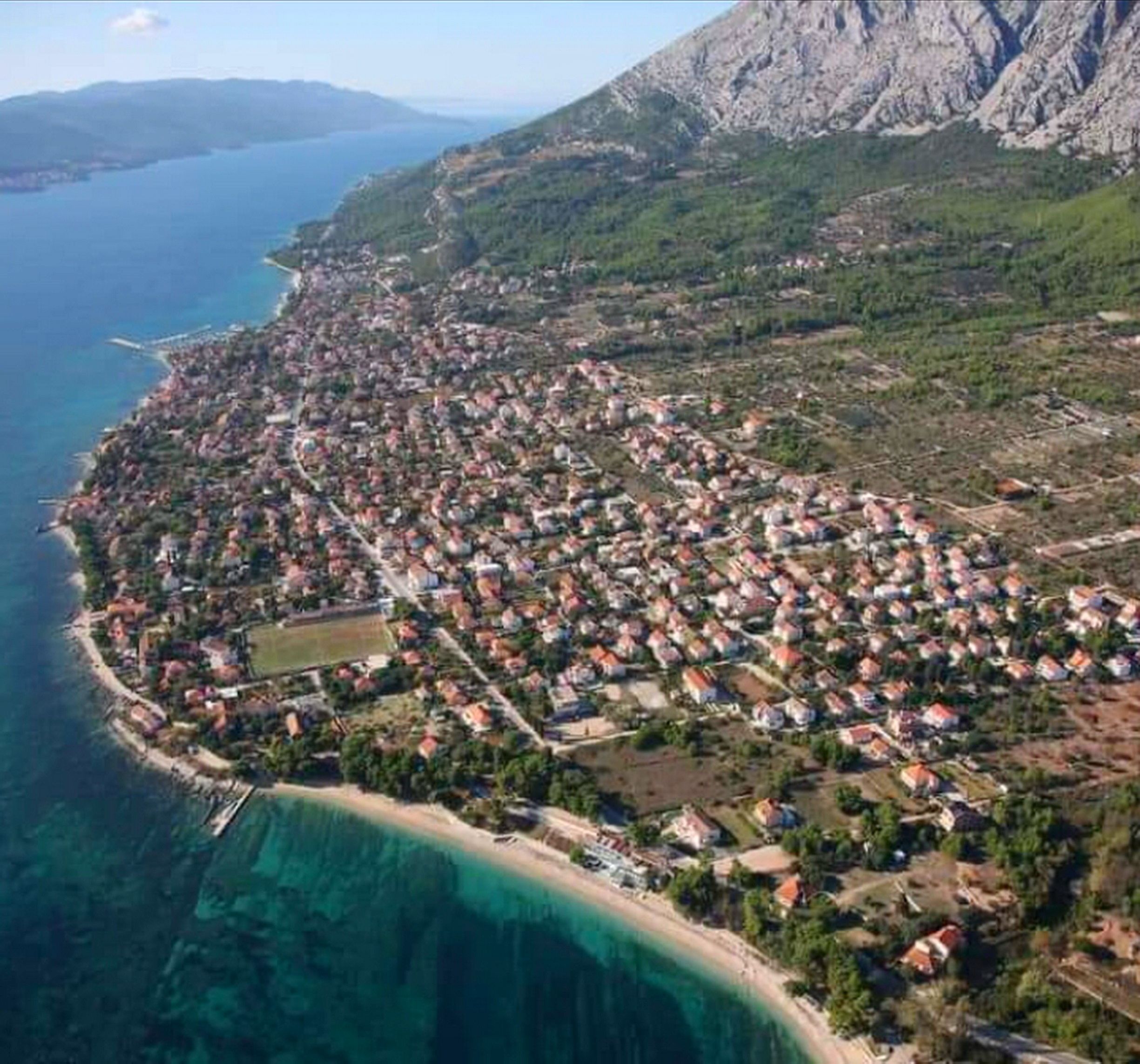 An aerial view of a coastal town with a dense cluster of houses, a clear coastline, and a mountainous backdrop.