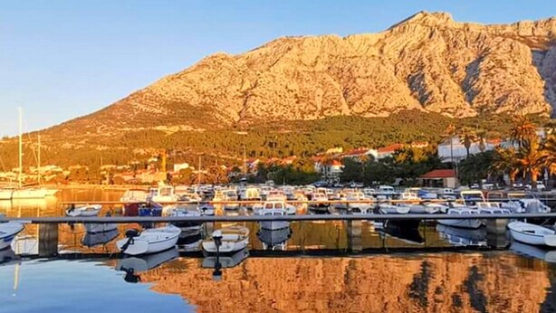 Una marina con barche ormeggiate in acque calme, una montagna sullo sfondo e cielo sereno.