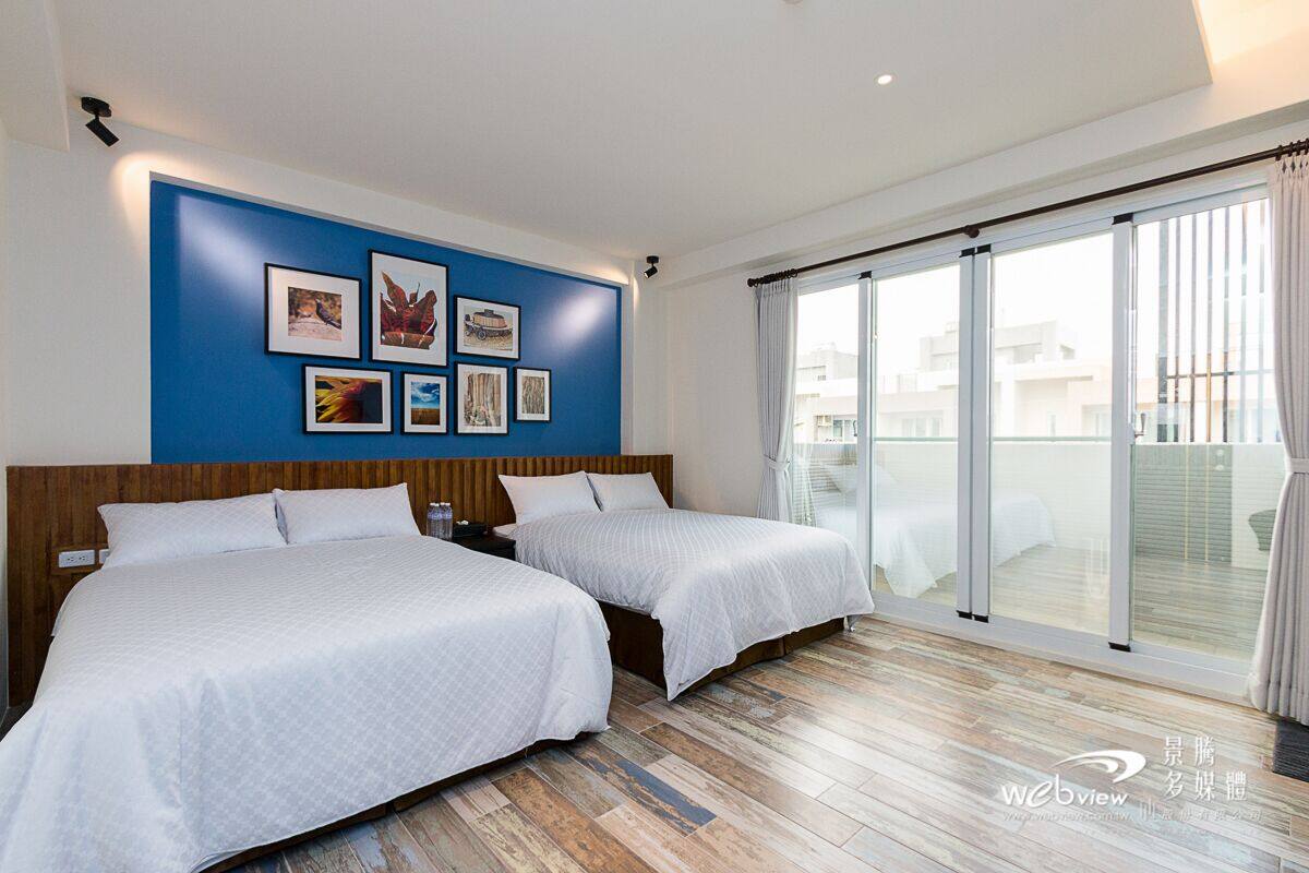 A hotel room with two beds, a blue accent wall with framed pictures, and a large sliding glass door to the balcony.