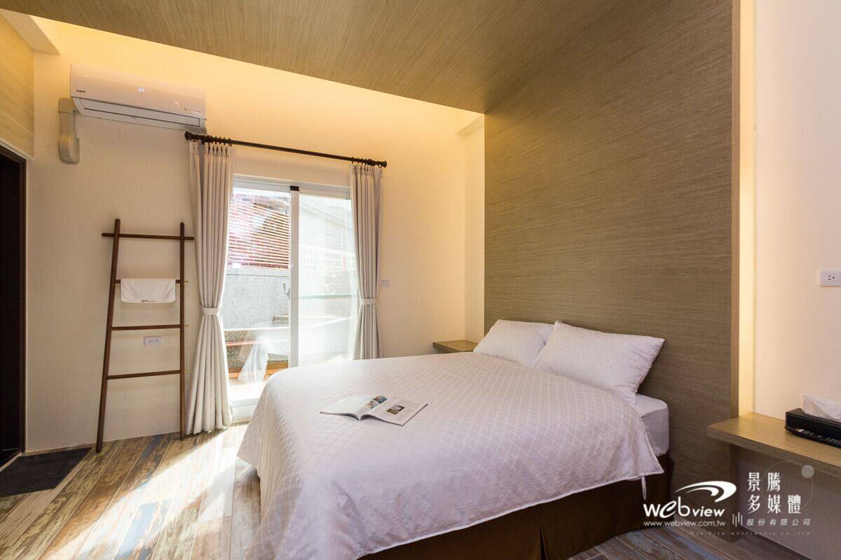 A hotel room with a bed, a nightstand, a window with curtains, a ladder, and an air conditioning unit.