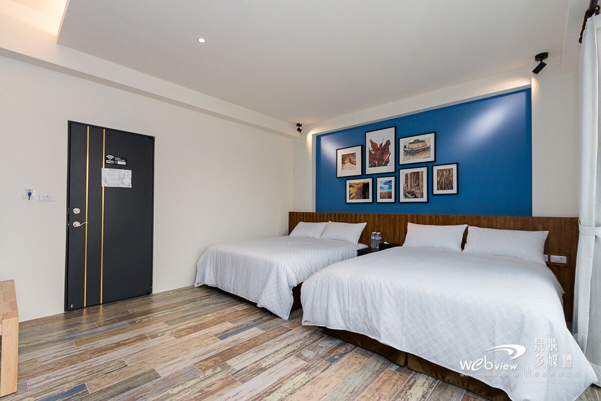 A hotel room with two beds, a blue accent wall with framed pictures, a wooden headboard, and a door with a sign.