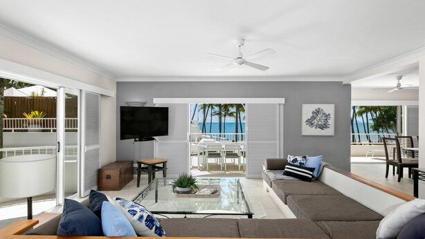 A modern living room with a large TV, a glass coffee table, and a sofa. There is a balcony with a view of the ocean and palm trees.