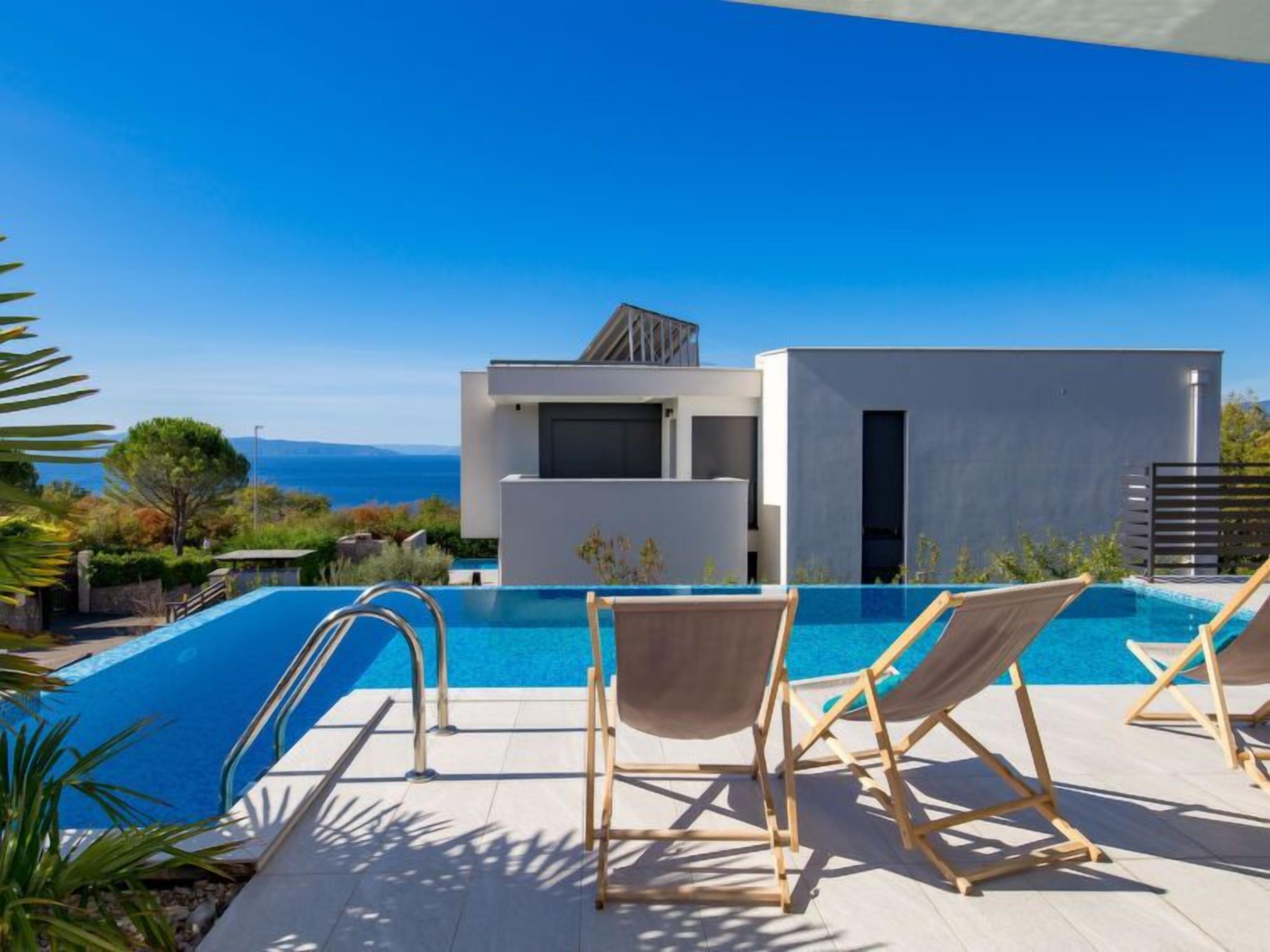 A modern pool area with a clear view of the sea, a contemporary building, and two wooden lounge chairs.
