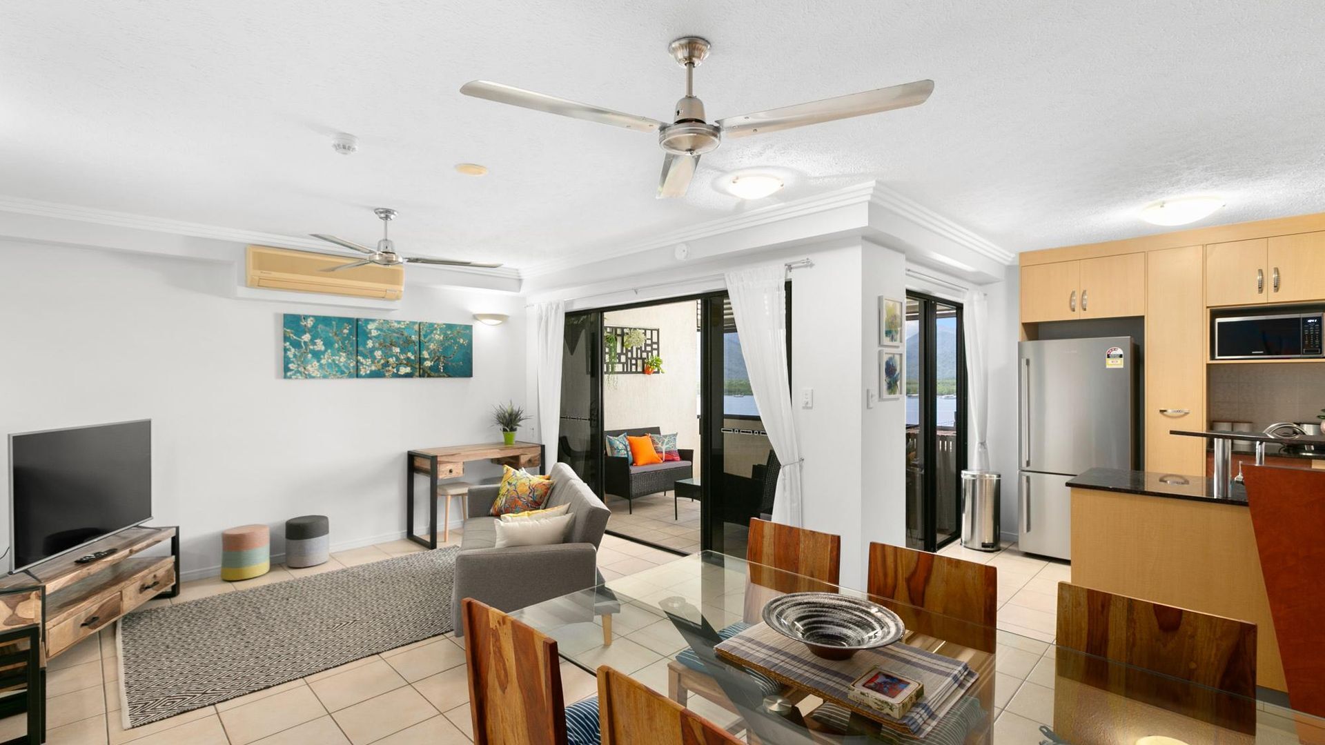 A modern living room with a TV, sofa, and dining area. There is a kitchen with cabinets and a refrigerator. A ceiling fan is visible, and the room has a large window.