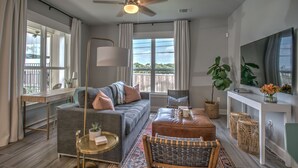 A modern living room with a grey sofa, a brown ottoman, a desk, a standing lamp, a television, and a balcony view. - Pool/Spa, guesthouse in the heart of ATX (Austin)