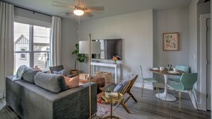 A modern living room with a grey sofa, a dining table with chairs, a ceiling fan, and a large window with curtains. - Pool/Spa, guesthouse in the heart of ATX (Austin)