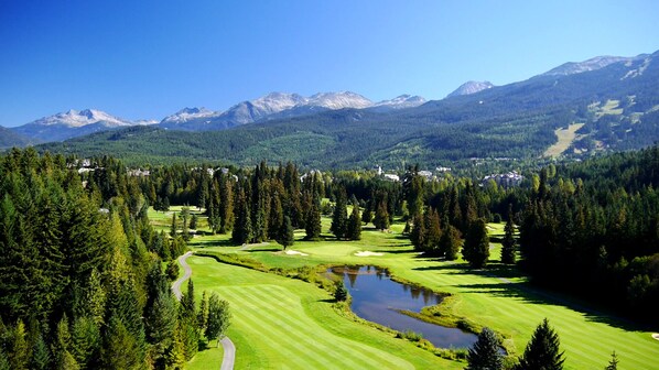 Golfkenttä, jonka yhteydessä on lampi, ja jota ympäröi tiheä metsä. Taustalla kohoavat vuoret.