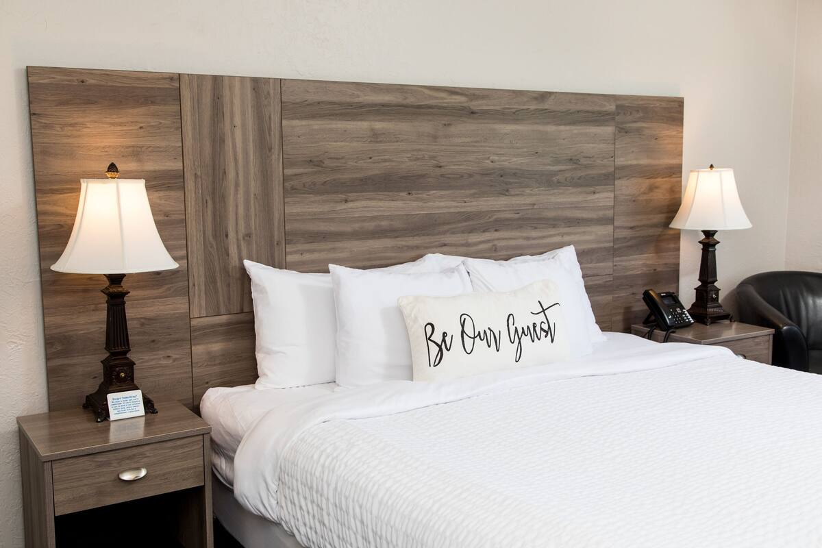 A neatly made bed with white bedding, two bedside lamps, a nightstand with a drawer, and a pillow with the text 'Be Our Guest.'