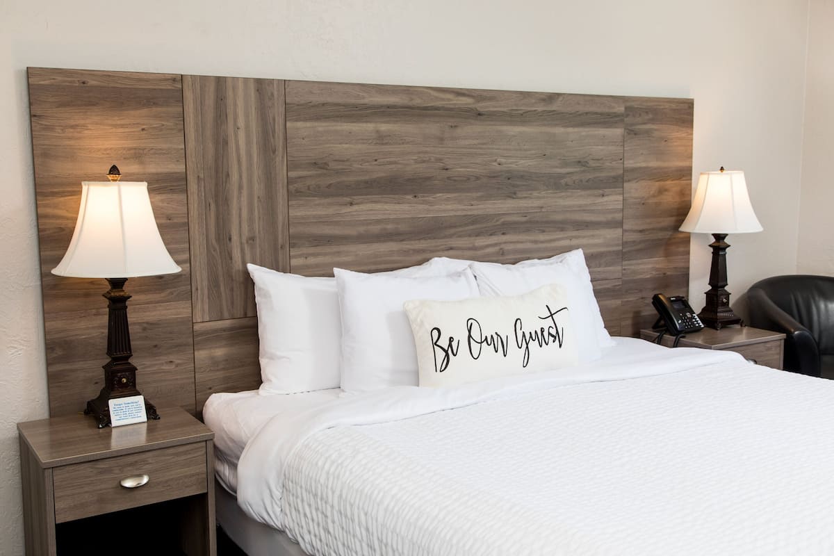A neatly made bed with white bedding and a pillow with the text "Be Our Guest." There are two bedside lamps on wooden nightstands, one on each side of the bed.