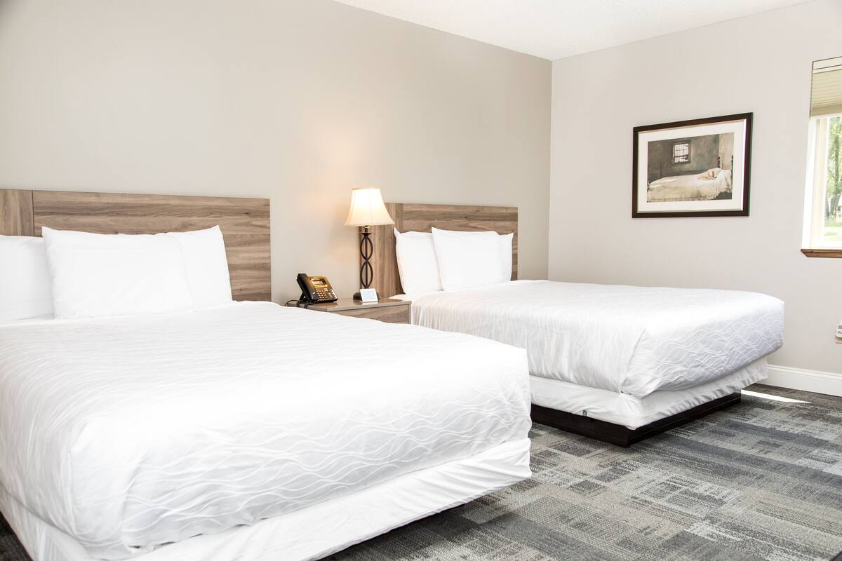 A hotel room with two beds, a nightstand with a lamp, a framed picture on the wall, and a window with blinds.