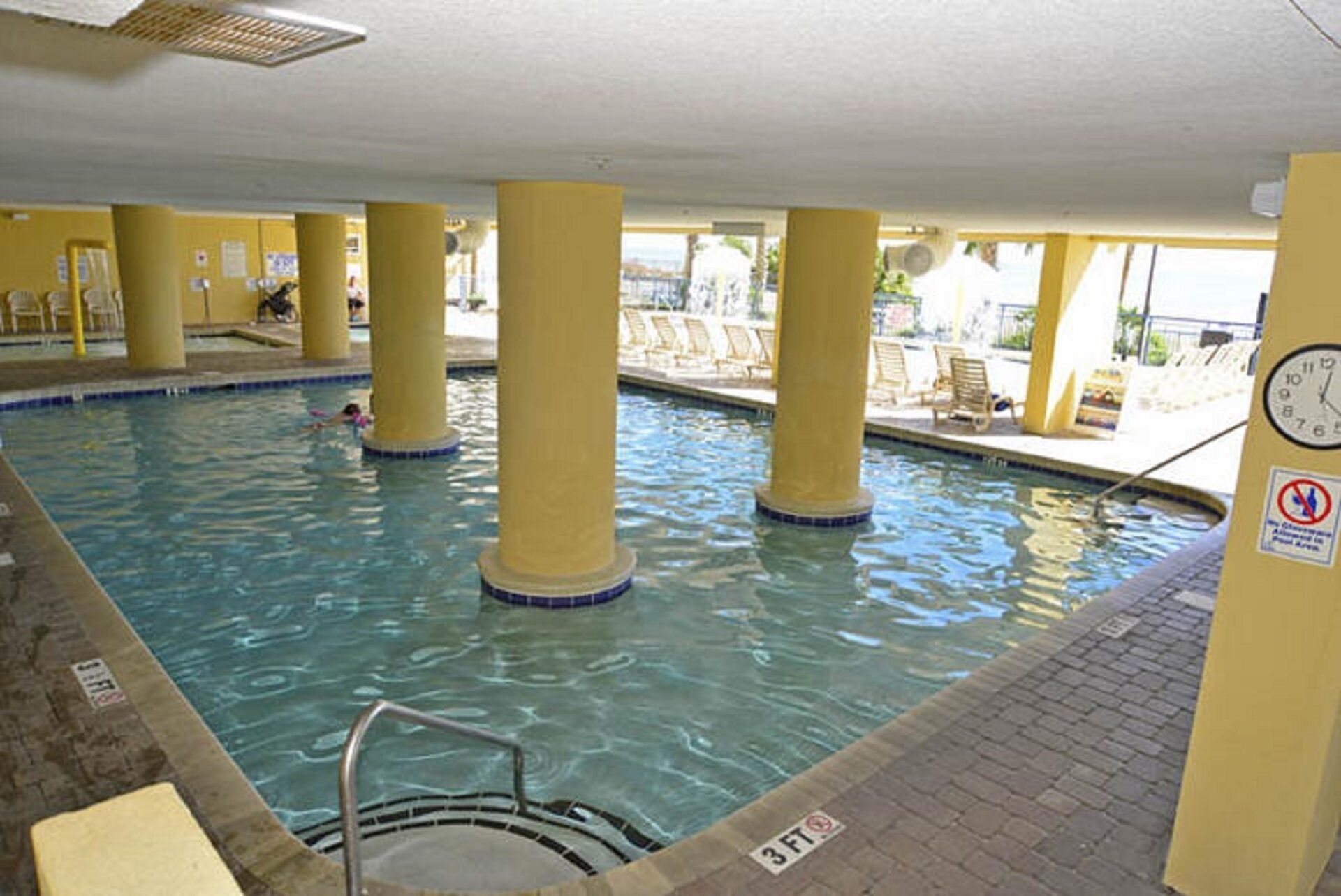 An indoor swimming pool with clear water, yellow pillars, a clock, and a no diving sign.