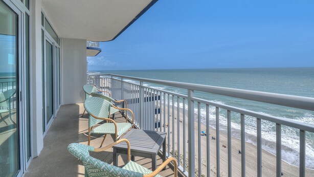 Un balcon avec des fauteuils en osier et une table, d’où l’on peut admirer une plage animée et un poste de surveillance des sauveteurs.