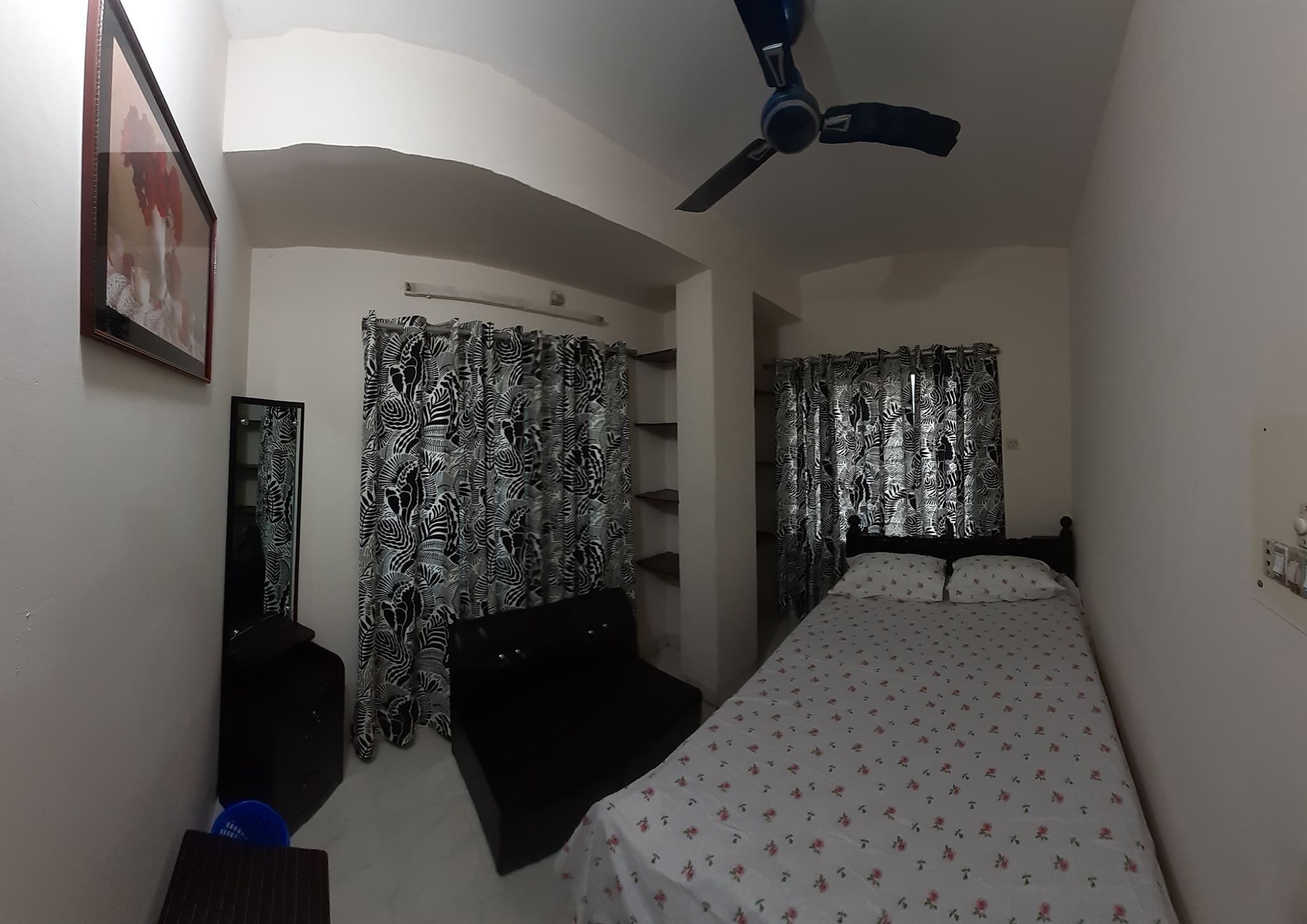 A bedroom with a bed, a ceiling fan, patterned curtains, a mirror, and a shelf.