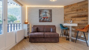 Un salon moderne avec un canapé en cuir marron, un mur en bois, un tableau représentant un téléphérique et un coin repas avec une table blanche et deux chaises.
