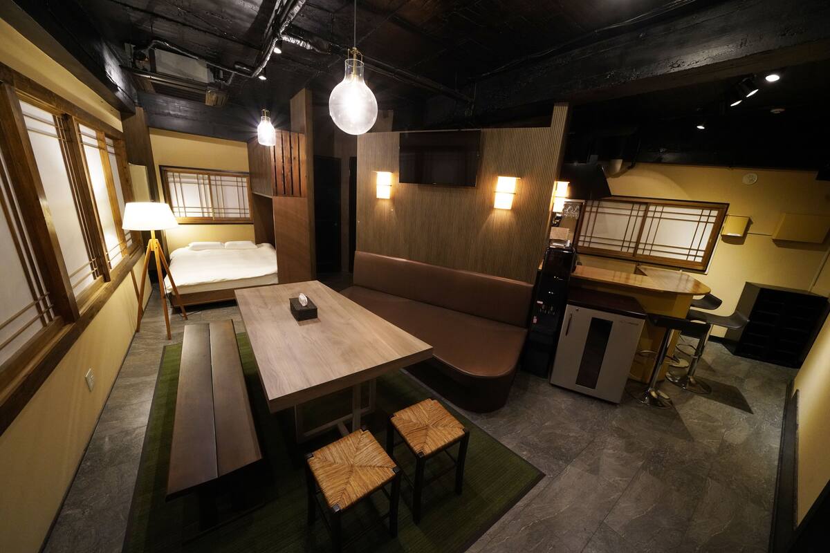 Habitación de hotel compacta con una mesa de comedor de madera, asientos tipo banco, una pequeña cocina americana y un televisor montado en la pared.