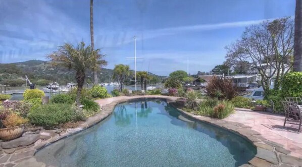 A swimming pool with a stone border, surrounded by landscaped greenery and a view of a marina with boats.