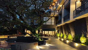 Un espacio exterior bien iluminado con un sendero curvo, áreas de descanso y un edificio con varios balcones y una pared de piedra.