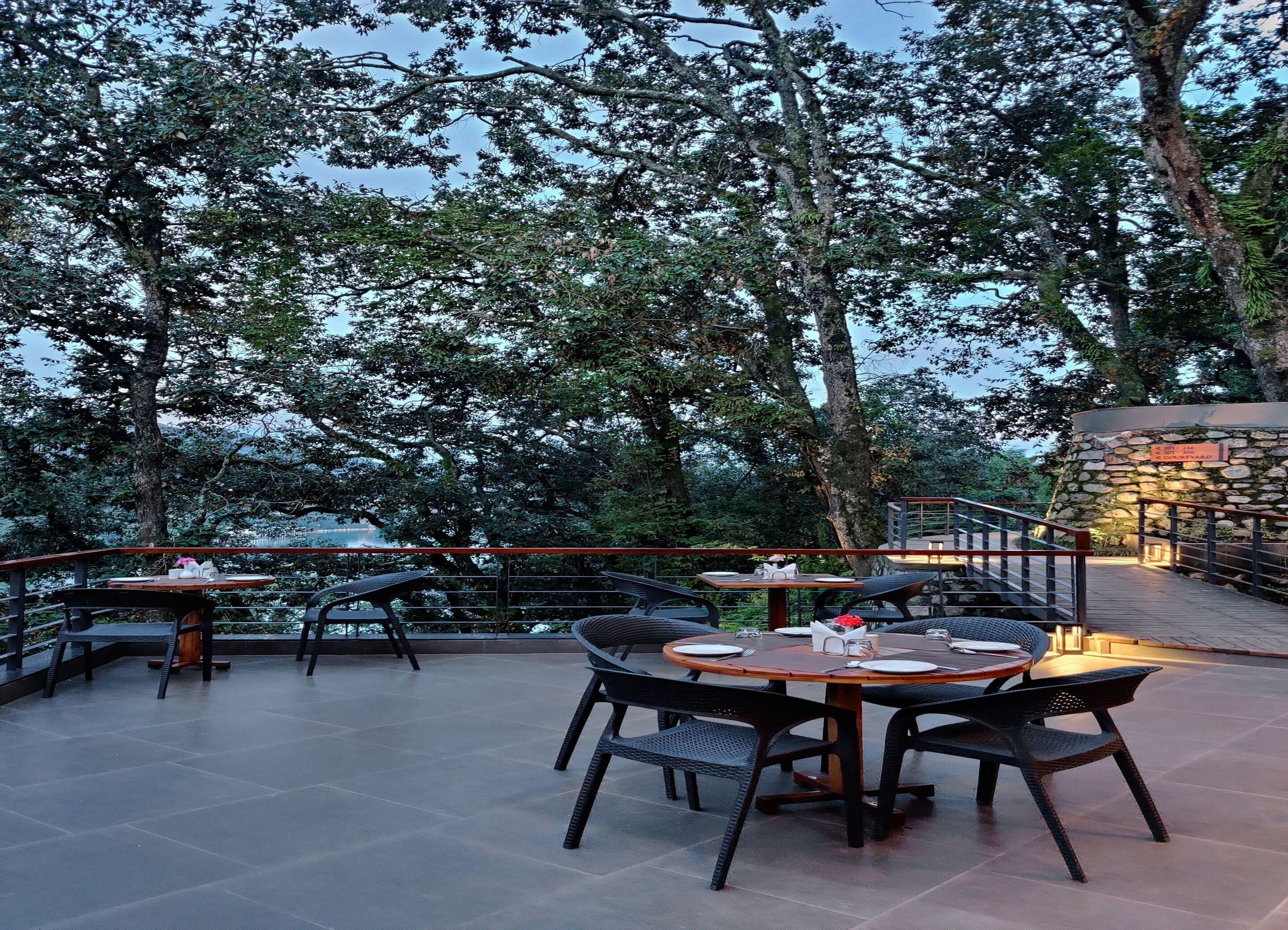 An outdoor dining area with tables and chairs, surrounded by trees and a stone wall.