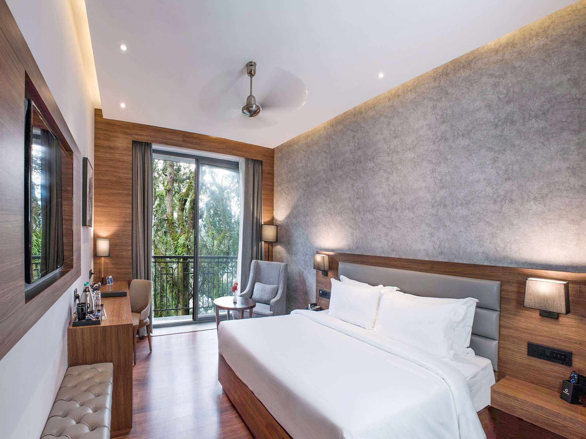 A modern hotel room with a large bed, a sitting area with a chair and table, and a balcony with a view of trees.