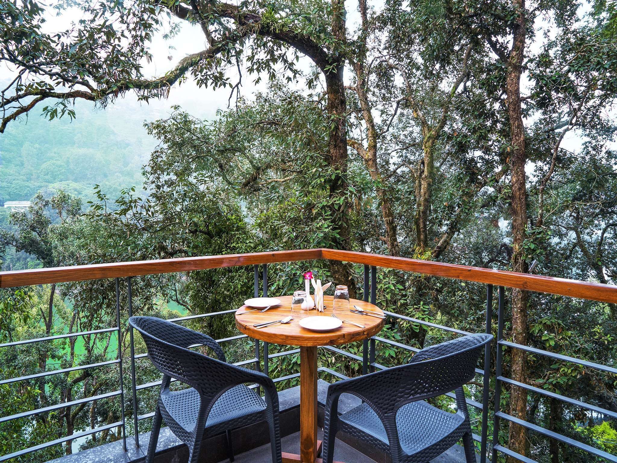 Une table entourée de chaises, placée sur un balcon délimité par une balustrade, d’où l’on peut admirer une zone boisée.