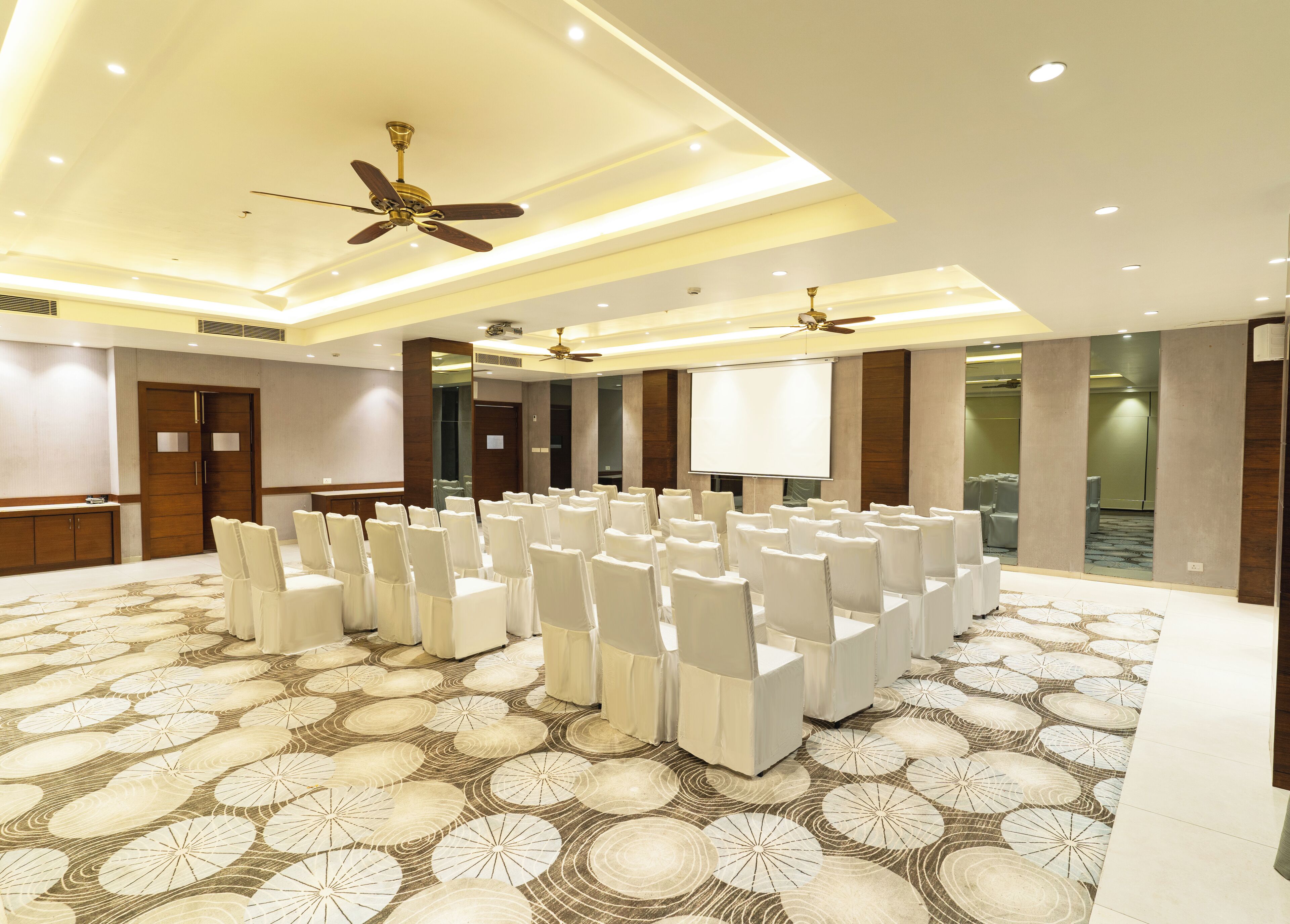 A conference room with rows of white chairs, a large screen, and ceiling fans.