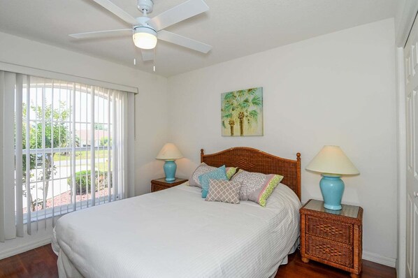 A bedroom with a bed, two nightstands, a ceiling fan, a window with blinds, and a painting on the wall.