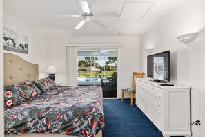 Hotelkamer met een bed, een bureau met een televisie, een stoel, een plafondventilator en uitzicht op een zwembad.