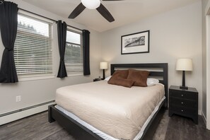 Une chambre à coucher avec un lit, deux tables de chevet avec des lampes, un ventilateur de plafond, une fenêtre avec des rideaux et un tableau encadré au mur.