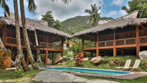 A pool area with two thatched-roof structures, surrounded by palm trees and large rocks. - Domaine Desaubin Luxury Villas (Mahé Island)