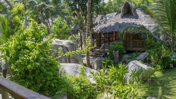 A rustic hut with a thatched roof surrounded by lush greenery and rocks. - Domaine Desaubin Luxury Villas (Mahé Island)