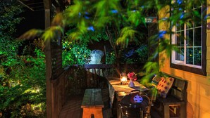 En udendørs terrasse med et træbord, stole, en lygte og en grill.