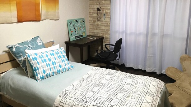 Ein Schlafzimmer mit einem Bett, zwei Kissen, einem Nachttisch, einem Sessel, einem Fenster mit Vorhängen und einer Klimaanlage.