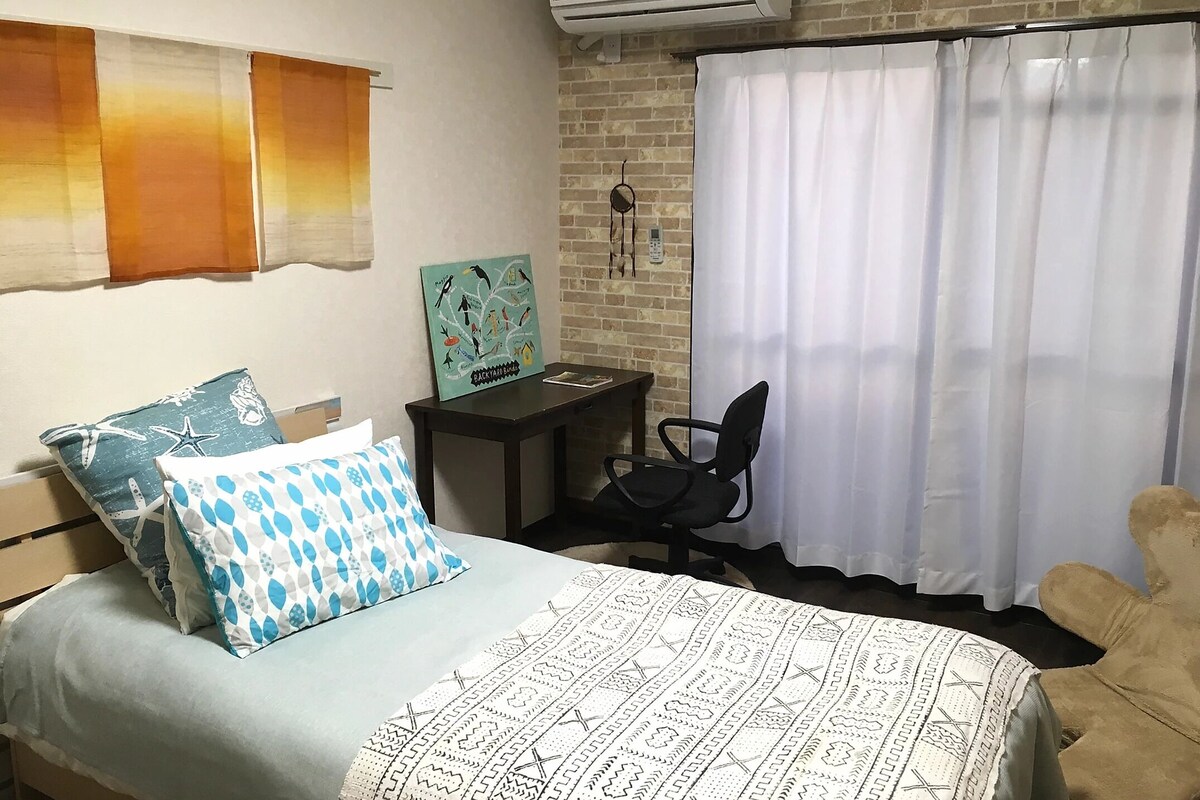 A bedroom with a bed, two pillows, a nightstand, a chair, a window with curtains, and an air conditioning unit.
