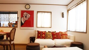 Een kamer met een eettafel, twee stoelen, een bank met gekleurde kussens, een klok aan de muur en een raam met gordijnen.