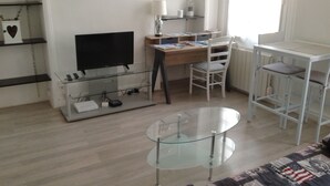 A living room with a glass TV stand, a wooden desk, a white chair, a glass coffee table, and a white radiator. - STUDIO by the water with all comfort (Le Havre)