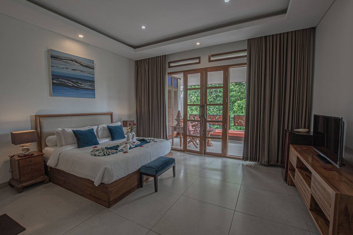 A bedroom with a bed, bedside table, bench, TV stand, and a view of the outdoors.