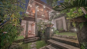 Una casa moderna con una escalera de piedra que lleva a una terraza de madera, rodeada de exuberante vegetación y un jardín cuidado.