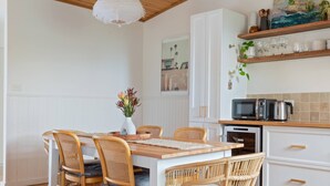 Een eethoek met een houten tafel en rieten stoelen, een witte vaas met bloemen, en een keuken met witte kasten en een magnetron.