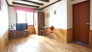 Una habitación con piso de madera, un televisor sobre un mueble de madera, dos sillas metálicas con cojines rojos, una mesita redonda y una puerta de madera.