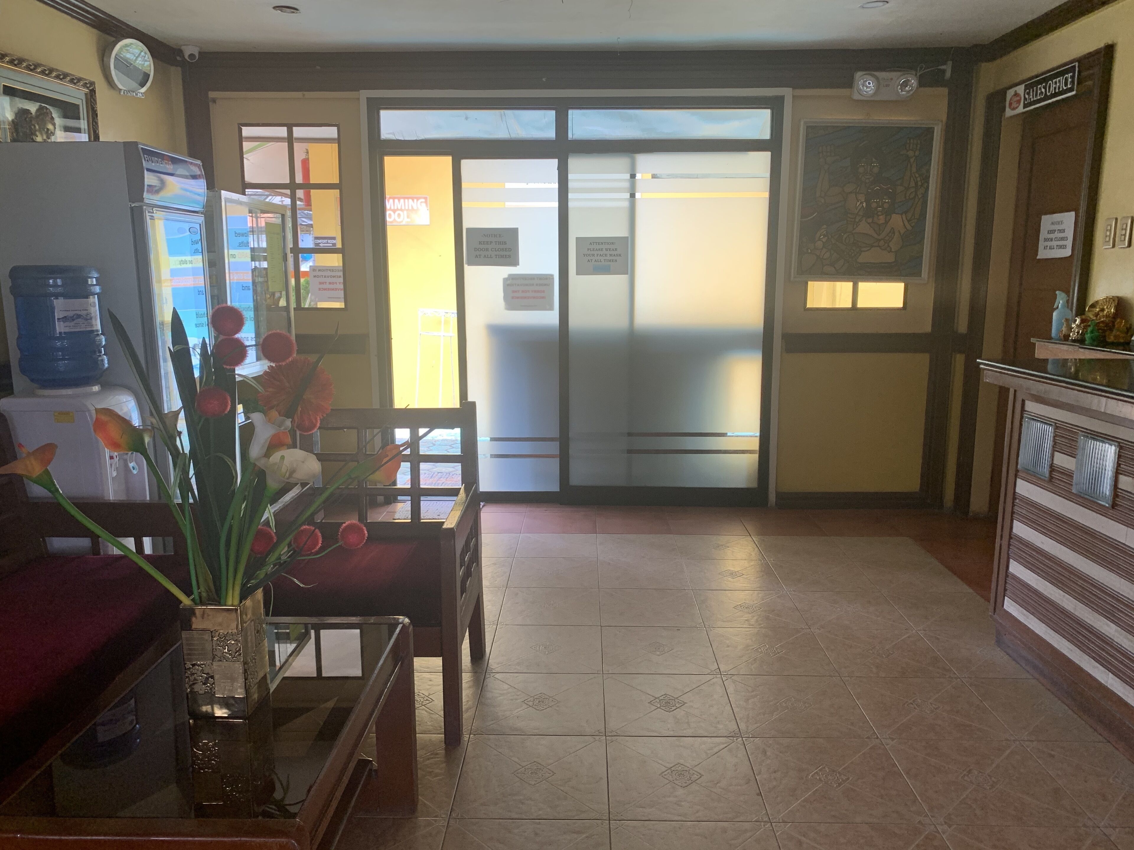 Zona de recepción con un enfriador de agua, un escritorio con un florero y una puerta con un letrero que dice 'Mantenga esta puerta cerrada en todo momento'.
