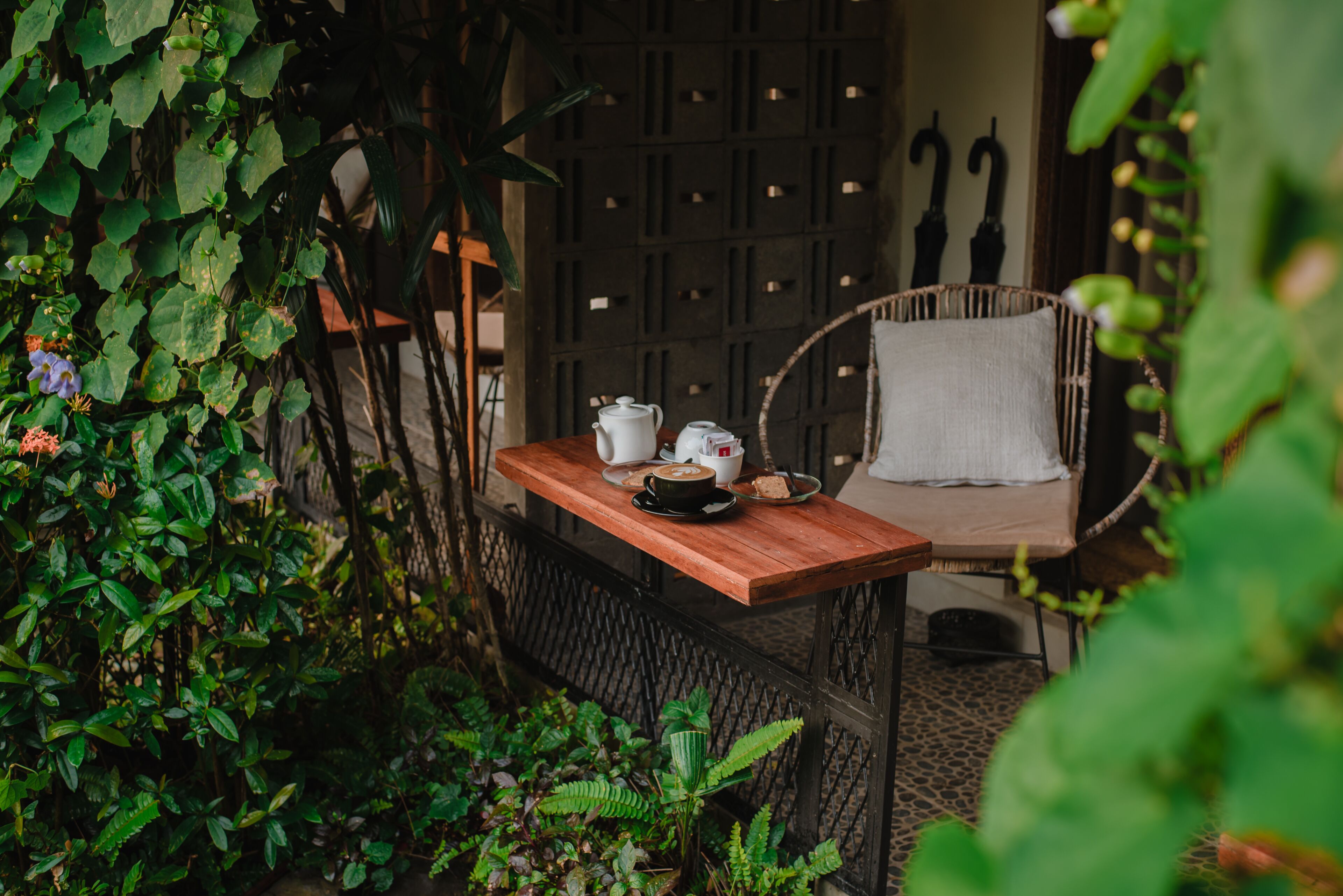 A wooden table with a white pitcher, a cup, and a plate with food, set against a wicker chair with a white cushion, surrounded by lush greenery.