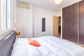 Ein ordentlich bezogenes Bett mit weißer Decke und einem einzelnen orangenen Kissen, eine Wand-Klimaanlage, ein gerahmtes Bild und eine Schiebetür zum Badezimmer.